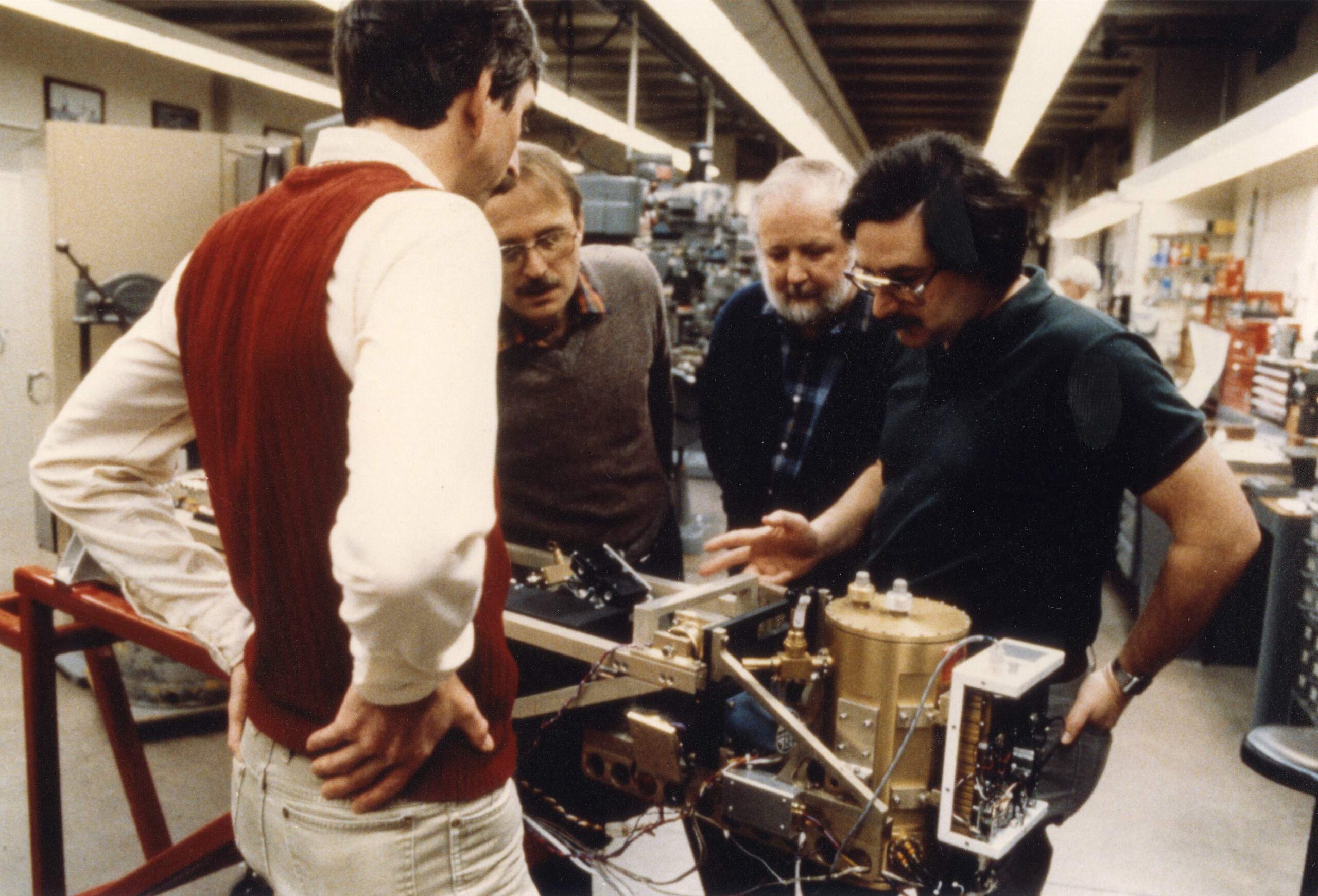 Four men inspect a small meteorological object consisting of metal canisters and electronics connected by wires.