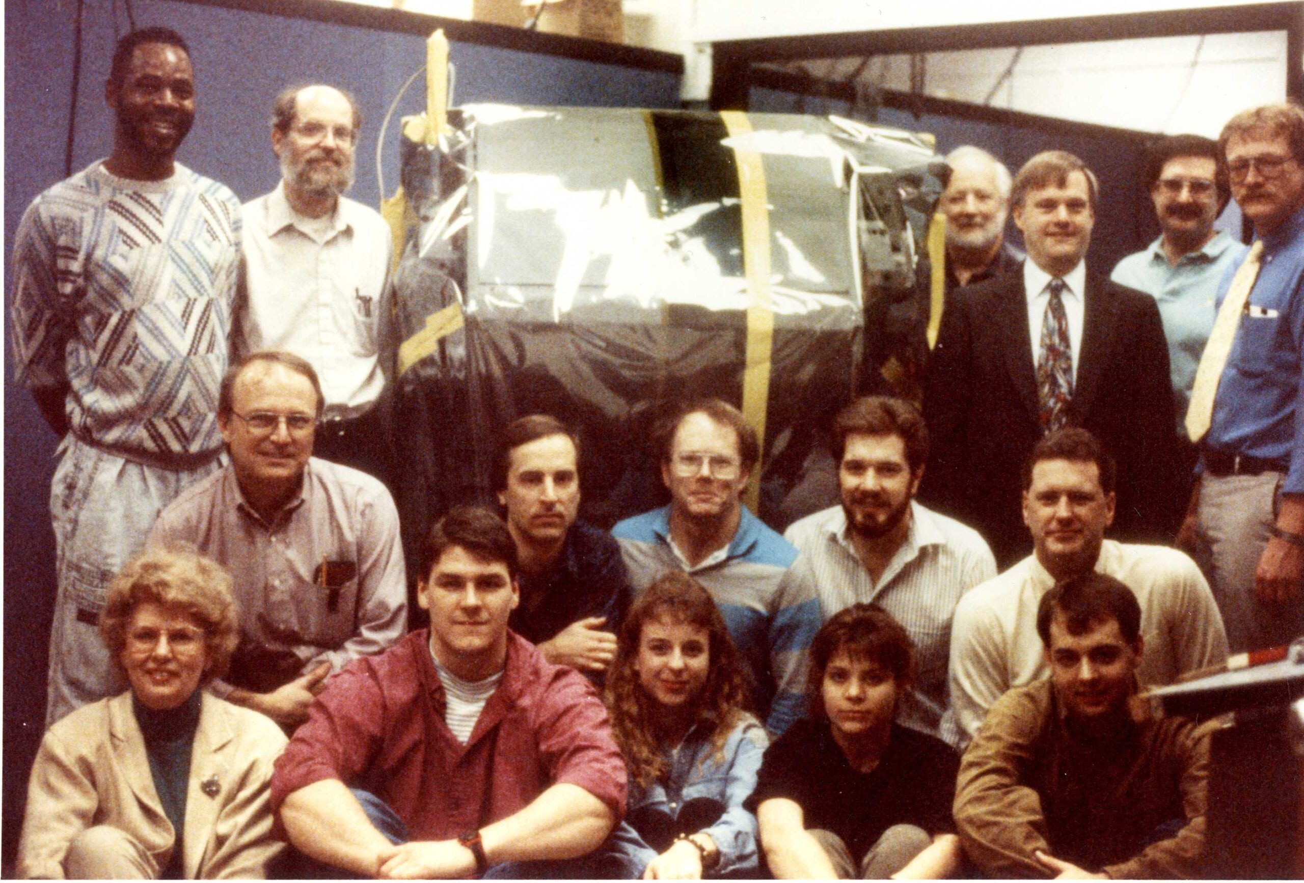 Sixteen people sit and stand around a large, cylindrical instrument wrapped in silvery foil.