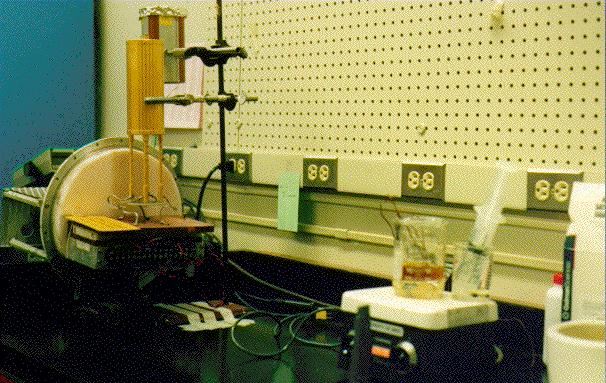 A gold canister about the size of a soda can is held upright by a stand on a laboratory table, surrounded by beakers, syringes, and electronic equipment.