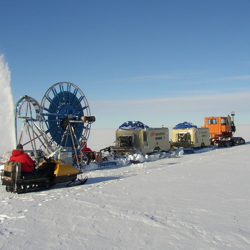 A drilling team on snowmobiles works in the middle of an ice field, operating a large metal wheel which turns and cranks the drill into the ice. 