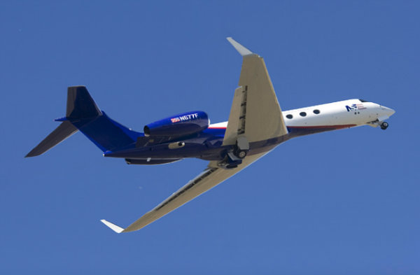 A small, sleek airplane flies up into a clear blue sky at a steep angle. The digits "M677F" are written on the engine.