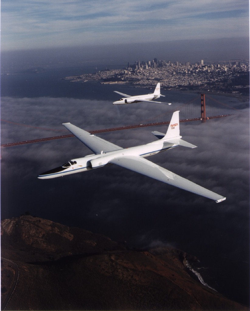 Two narrow airplanes with long, rounded noses fly low above the Golden Gate Bridge on a partly cloudy day. 