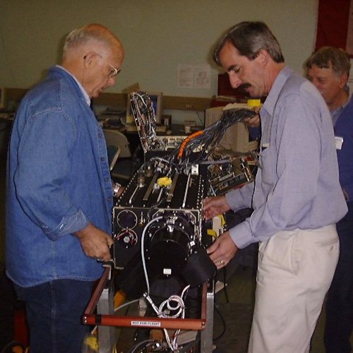 Three men wearing casual dress clothes work on a mid-sized, boxy, complicated piece of equipment with knobs, wires, and sensors.