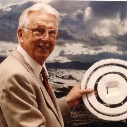 Verner Suomi, wearing a tan suit, glasses, and a red tie, stands before a choppy ocean scene holding a circular disk made of foam. 