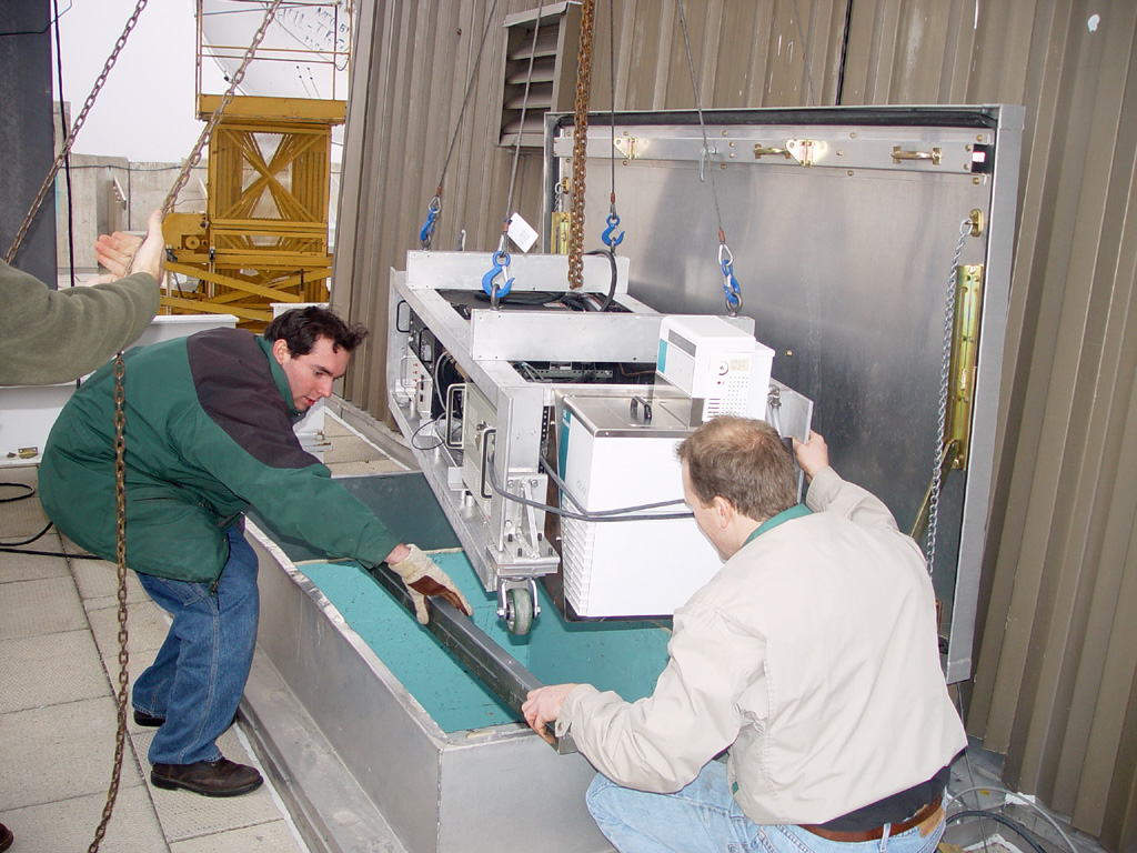 Three people wearing coats work to hoist a fridge-sized white box containing the AHSRL laser water chiller and lidar electronics onto the roof of the AOSS Building.
