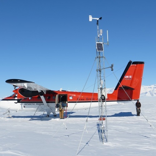 A red and white small airplane sits on a snowfield under a clear blue sky. Written on the side of the airplane are the words "US Antarctic Program". A person stands at the open door of the airplane, looking in. In the foreground stands a short metal tower with various meteorology instruments and a solar panel attached to it. 
