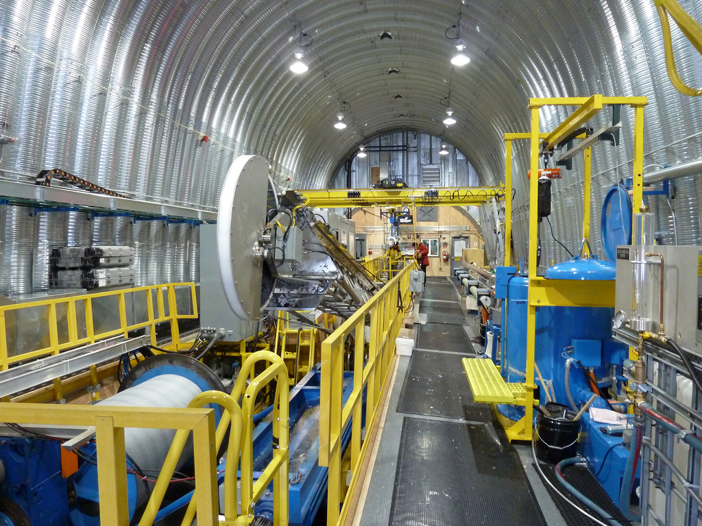 Ice drilling equipment painted bright blue and yellow sits inside a long and narrow warehouse. 