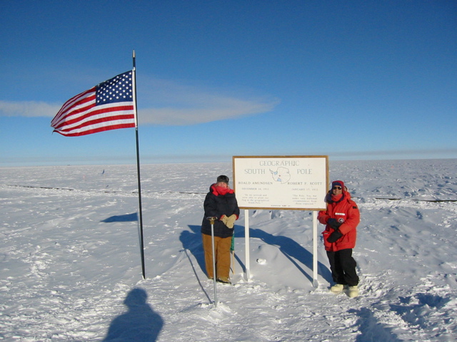 Two people wearing heavy snow gear stand in the middle of an ice field next to a flagpole flying an American flag and a large sign reading "Geographic South Pole".