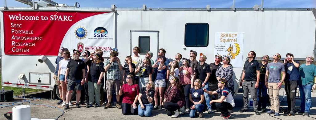 A large group of people stand next to a white trailer, wearing eclipse glasses and looking up at the sun. The trailer sports two banners: one reading "Welcome to SPARC: SSEC Portable Atmospheric Research Center", the other reading "Sparcy Squirrel" with a cartoon drawing of a yellow squirrel. 