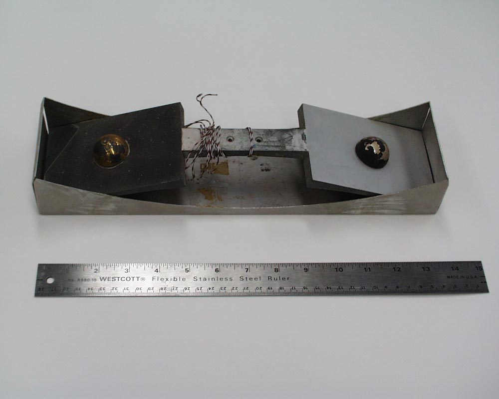A flat, metal instrument 13.5 inches in length with two wide, flat paddles of metal on both ends. Centered on each paddle is a raised metal hemisphere.
